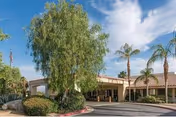 Exterior view of ManorCare Health Services-Palm Desert facility showing the entrance with a covered drop-off area, surrounded by palm trees, bushes, and a clear blue sky with some clouds.