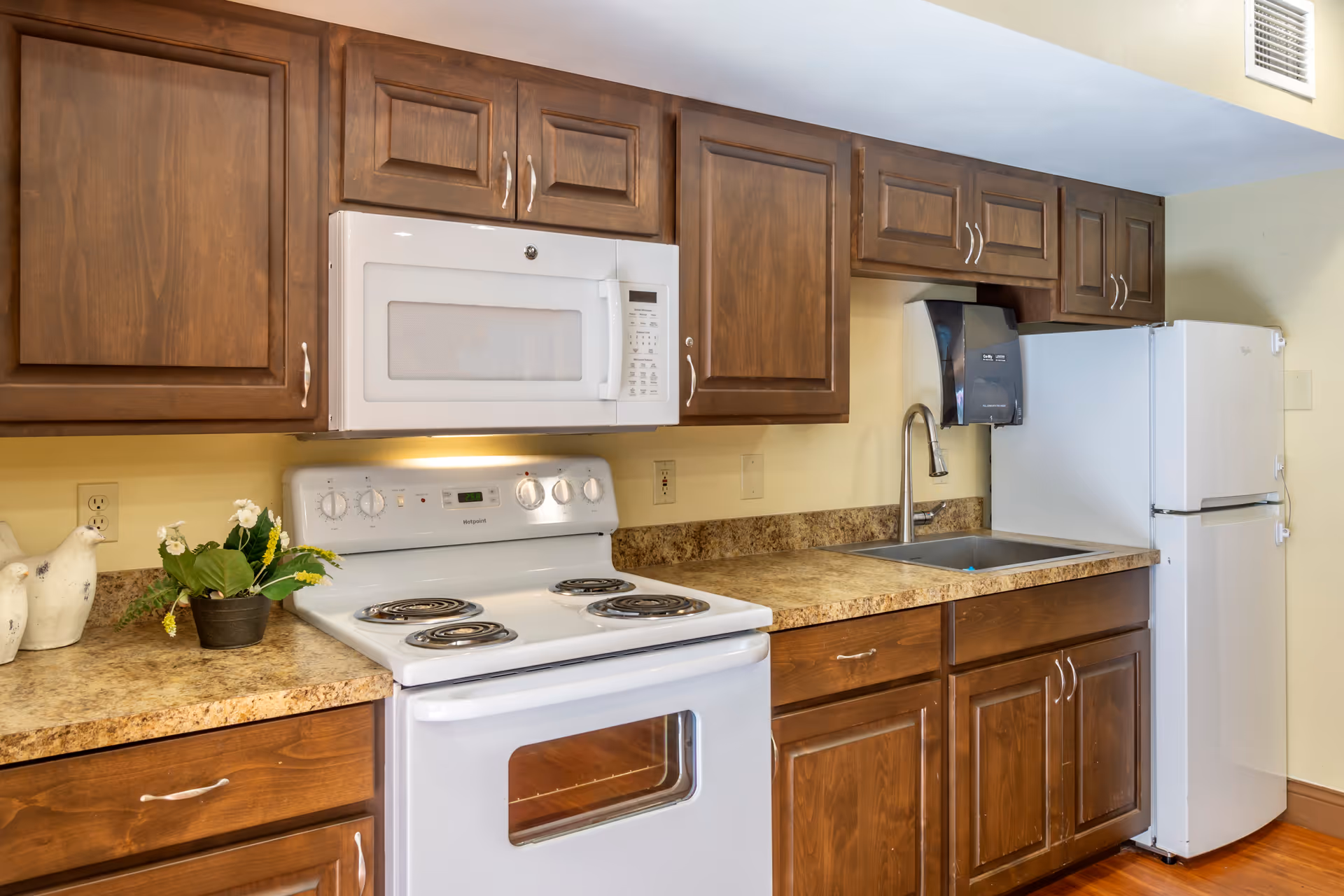 Compact kitchen with wooden cabinets, a white stove and microwave, sink, refrigerator, and a small potted plant on the counter.