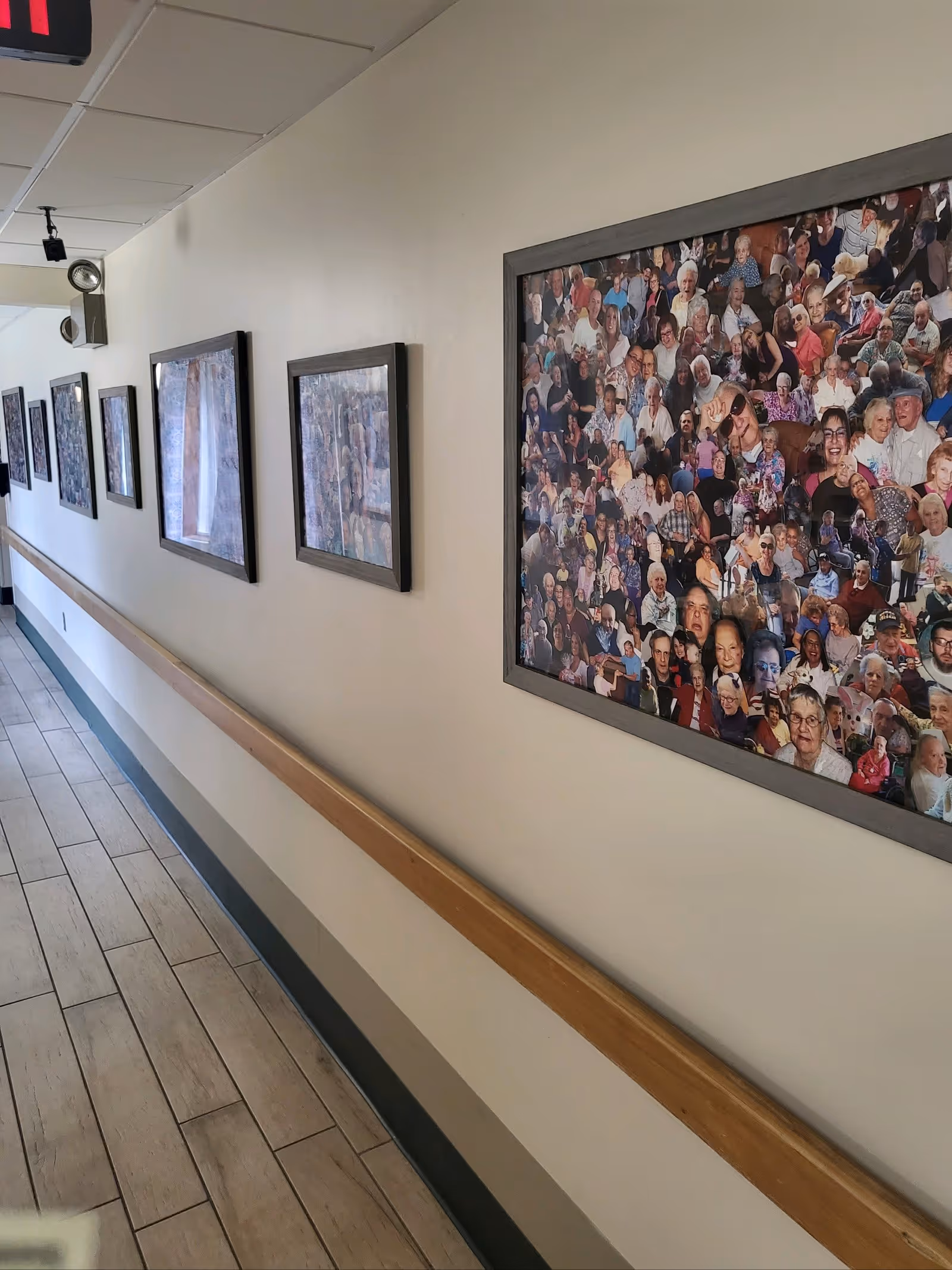 A hallway in a senior living facility with wooden handrails along the wall and several framed pictures hanging above the handrails. The floor is tiled with light-colored tiles, and the ceiling has recessed lighting. One of the pictures is a collage of many photos of elderly residents.