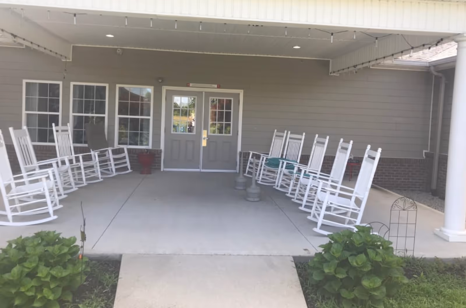 Covered outdoor porch area with white rocking chairs arranged in two rows facing each other, a concrete floor, and a double door entrance with windows on either side. There are green plants on either side of the walkway leading to the porch.