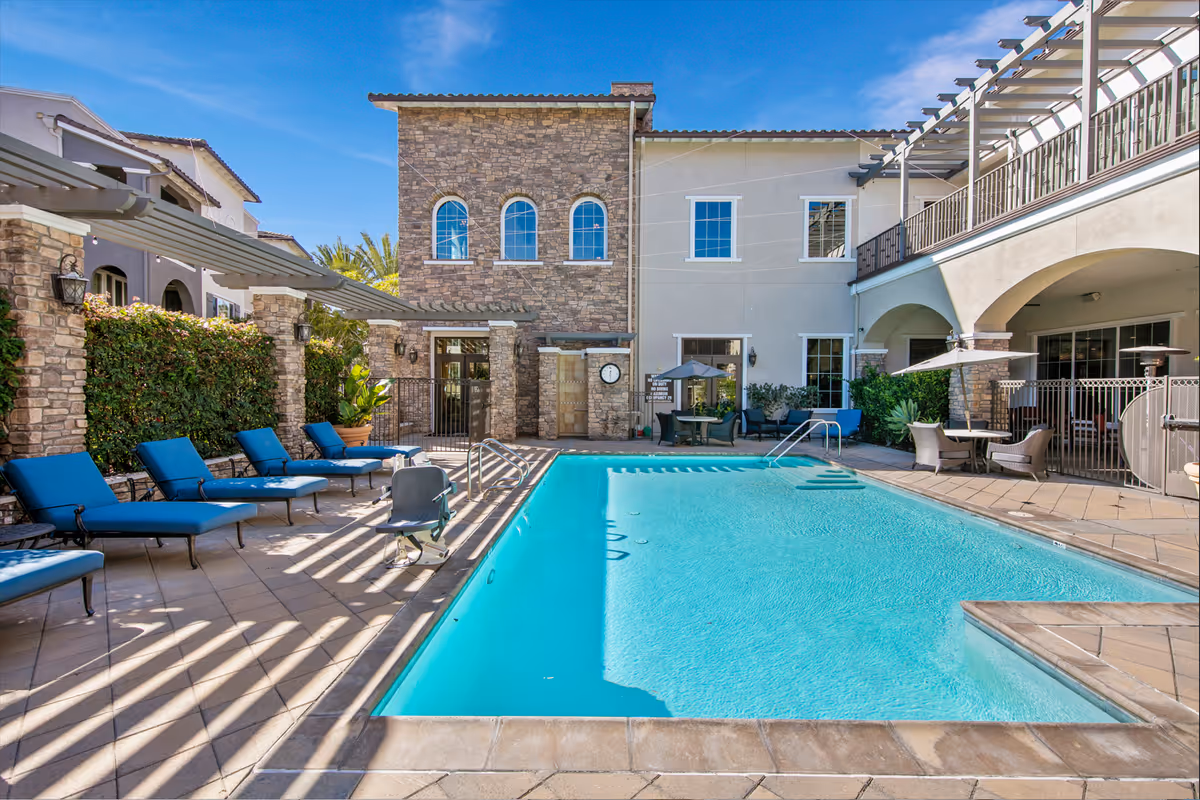 Outdoor swimming pool area at Meridian at Lake San Marcos with blue lounge chairs lined up on the left side, a pool lift for accessibility, and tables with umbrellas on the right side. The building surrounding the pool has stone and stucco walls with windows and balconies under a clear blue sky.