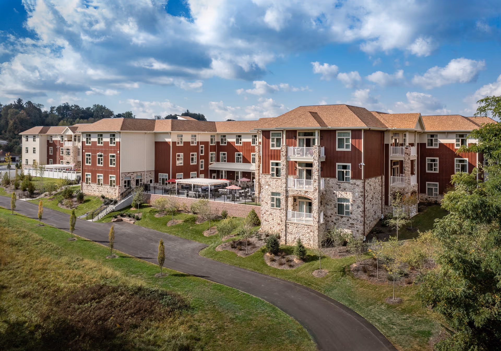 Exterior view of a large senior living facility building with a mix of stone and red siding, multiple windows, balconies, and a patio area with tables and umbrellas. The building is surrounded by landscaped greenery and a paved driveway under a partly cloudy blue sky.
