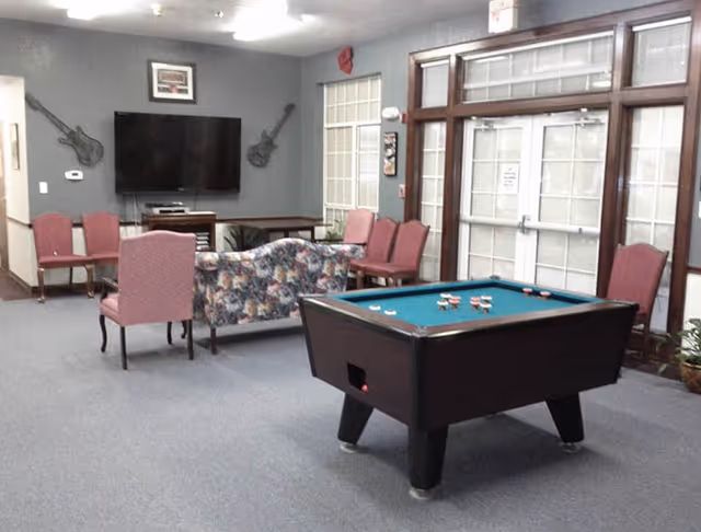 A common area in an assisted living facility featuring a pool table in the foreground, several chairs and a floral-patterned loveseat arranged facing a wall-mounted flat screen TV. The walls are decorated with two guitar-shaped wall hangings and a framed picture. Large windows with white blinds and a glass door are visible on the right side.