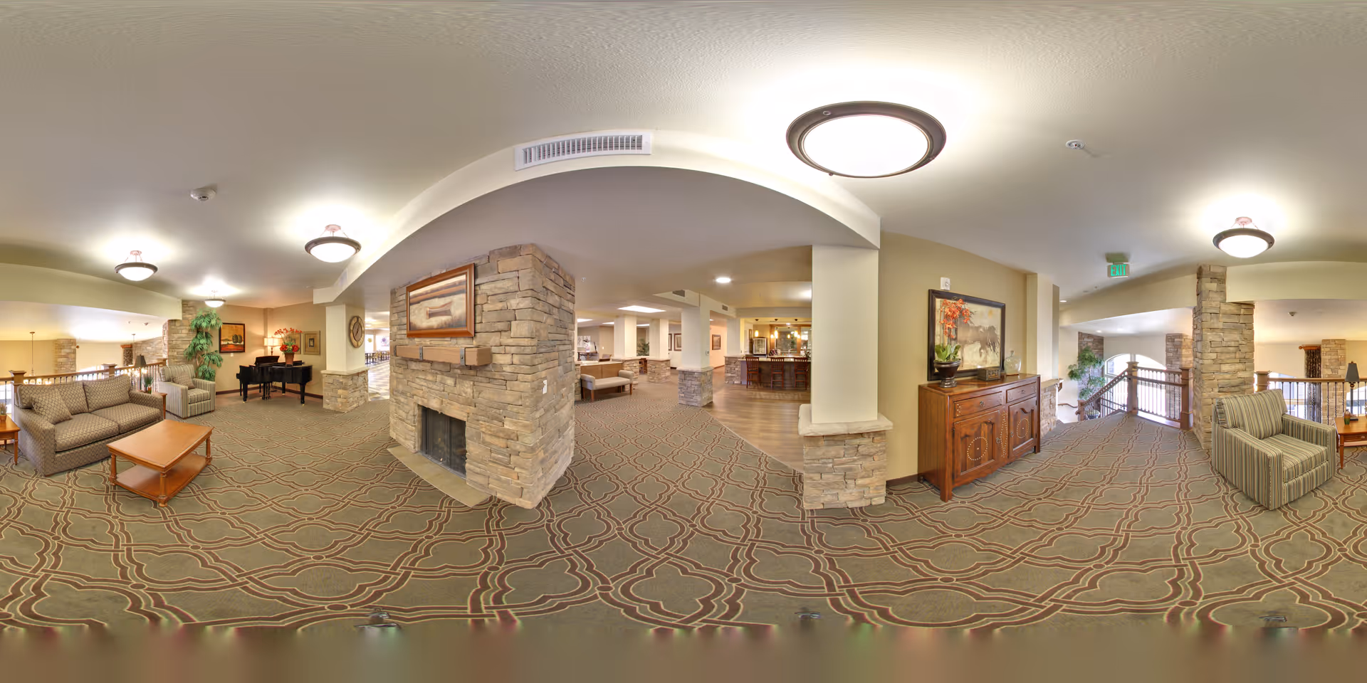 Spacious senior living facility common area with patterned carpet, stone fireplace, comfortable seating including sofas and armchairs, wooden tables, decorative plants, framed artwork, and warm lighting fixtures on the ceiling.