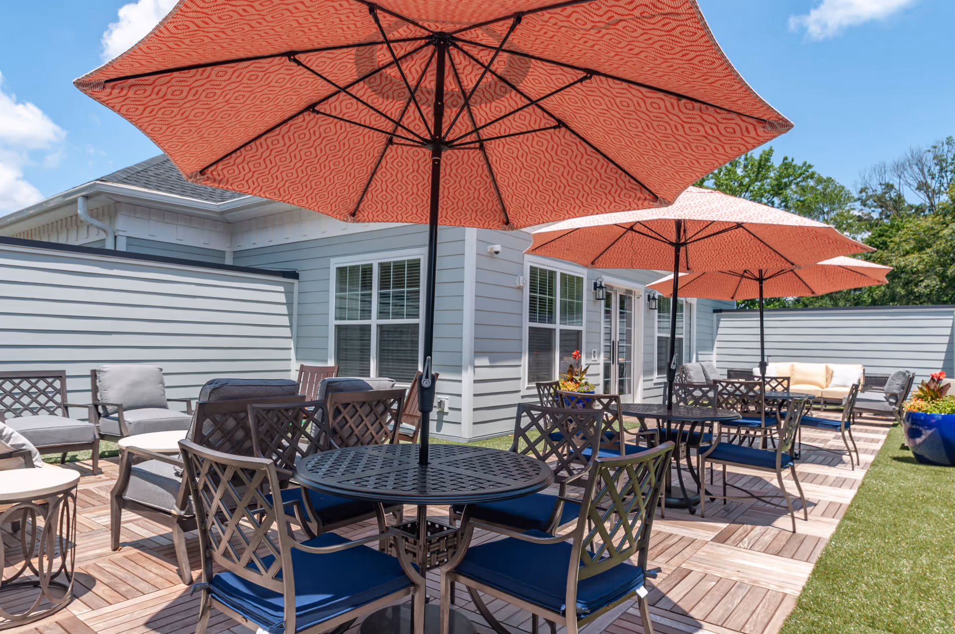 Outdoor patio area with multiple round tables and chairs, each shaded by large red patterned umbrellas. The patio has wooden flooring tiles and is adjacent to a light gray building with white trim and large windows. There are also cushioned lounge chairs and potted plants visible under a clear blue sky.