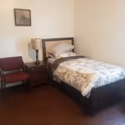 A small bedroom with a single bed featuring a patterned comforter and pillow. Next to the bed is a wooden nightstand with a lamp on it. A red cushioned chair is placed beside the nightstand. A framed picture hangs on the wall above the chair.