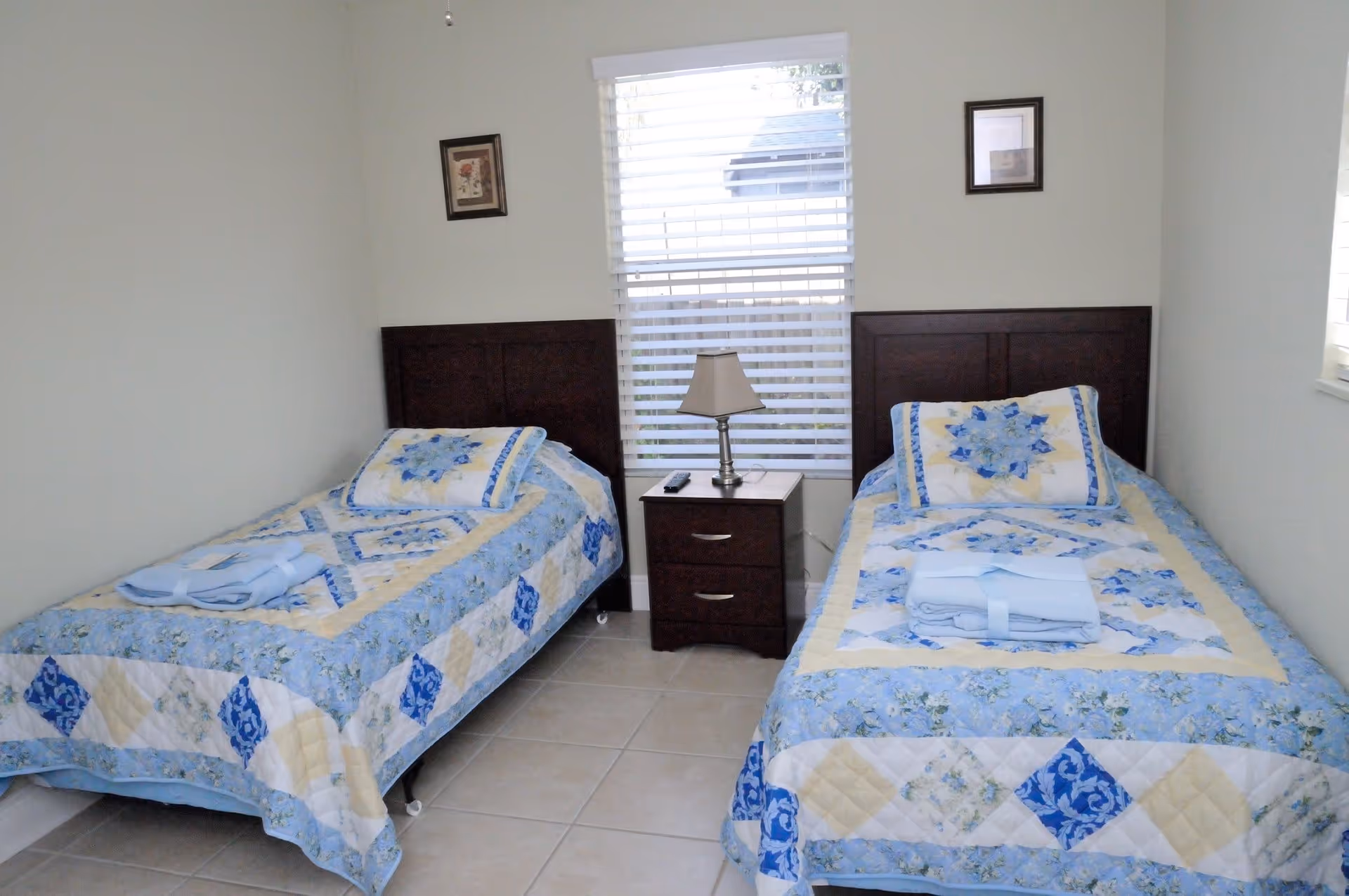 A small bedroom with two twin beds featuring blue, yellow, and white patterned quilts and matching pillows. Each bed has a folded towel set placed on top. Between the beds is a dark wooden nightstand with two drawers, a lamp, and a remote control. A window with white blinds is centered above the nightstand, and two framed pictures hang on the walls above each bed.