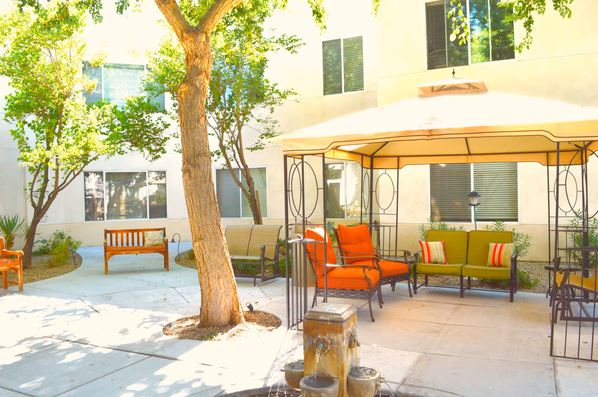 Outdoor courtyard area with a large tree, a gazebo with orange and green cushioned chairs, wooden benches, and a small water fountain in the foreground. The courtyard is surrounded by a building with several windows.