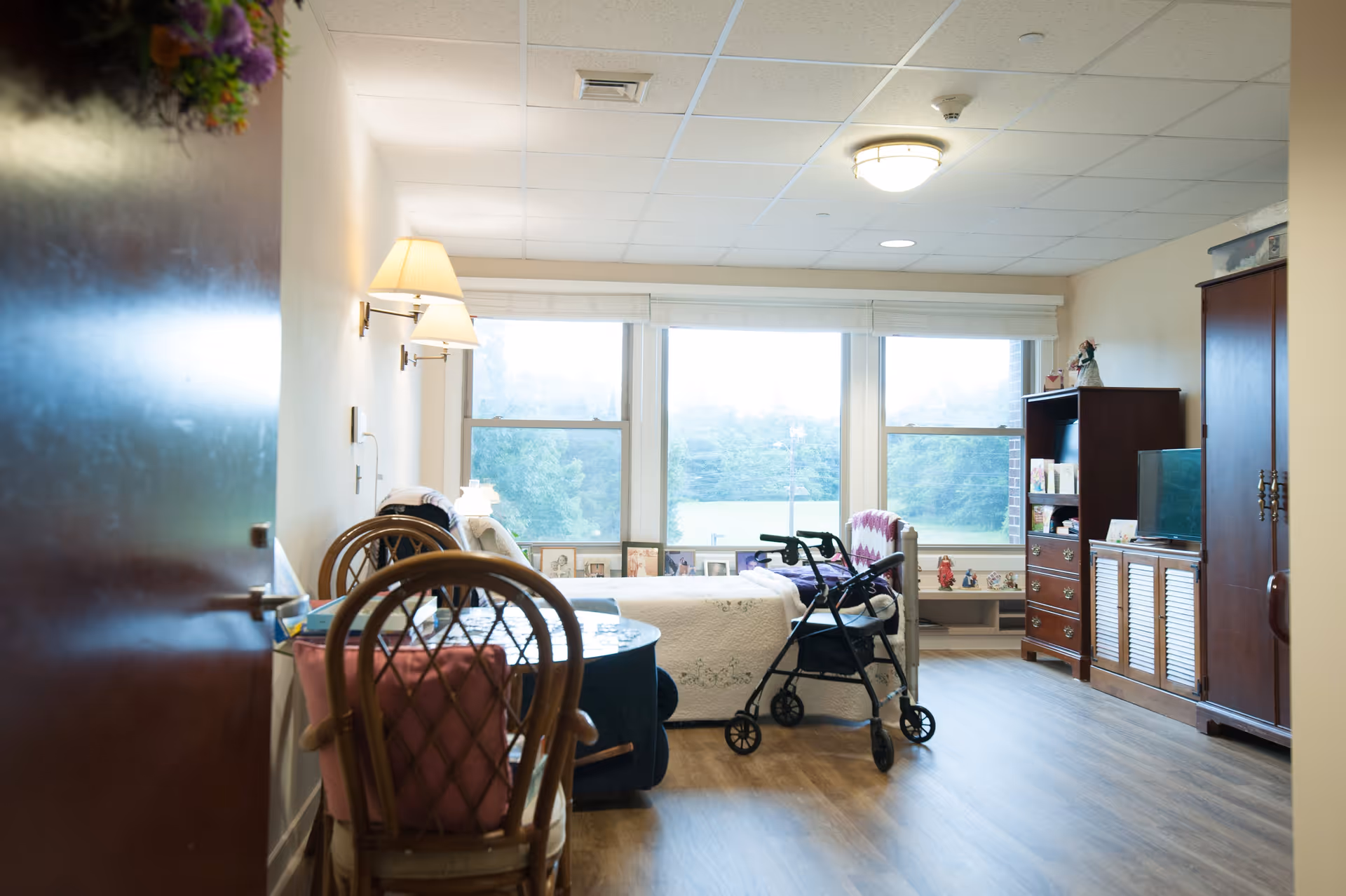 A bright and cozy senior living bedroom with a bed near large windows showing a green outdoor view. The room includes a walker, a wooden chair with a pink cushion, a small round table, a wooden dresser with a TV, and a wardrobe. The walls are light-colored, and the floor is wooden. There are framed photos on the windowsill and decorative items on the furniture.