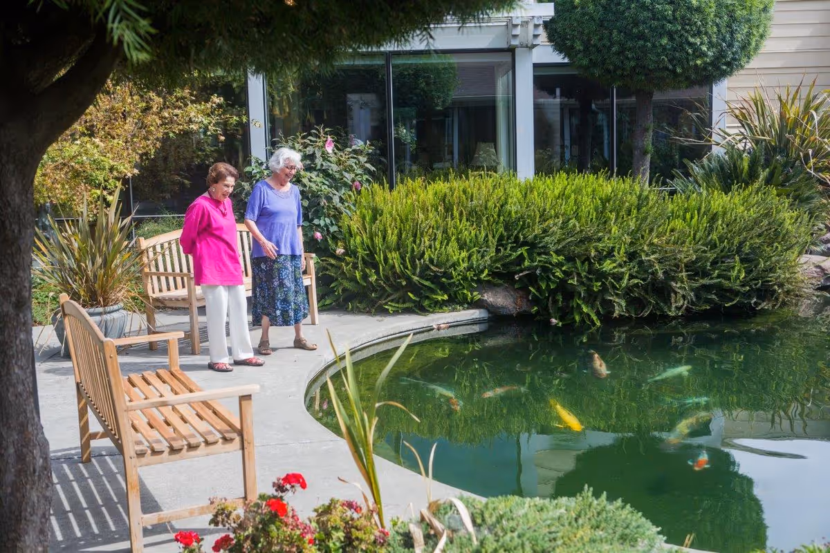 Two elderly women standing near a koi pond in a garden area outside a building, surrounded by greenery, flowers, and wooden benches.