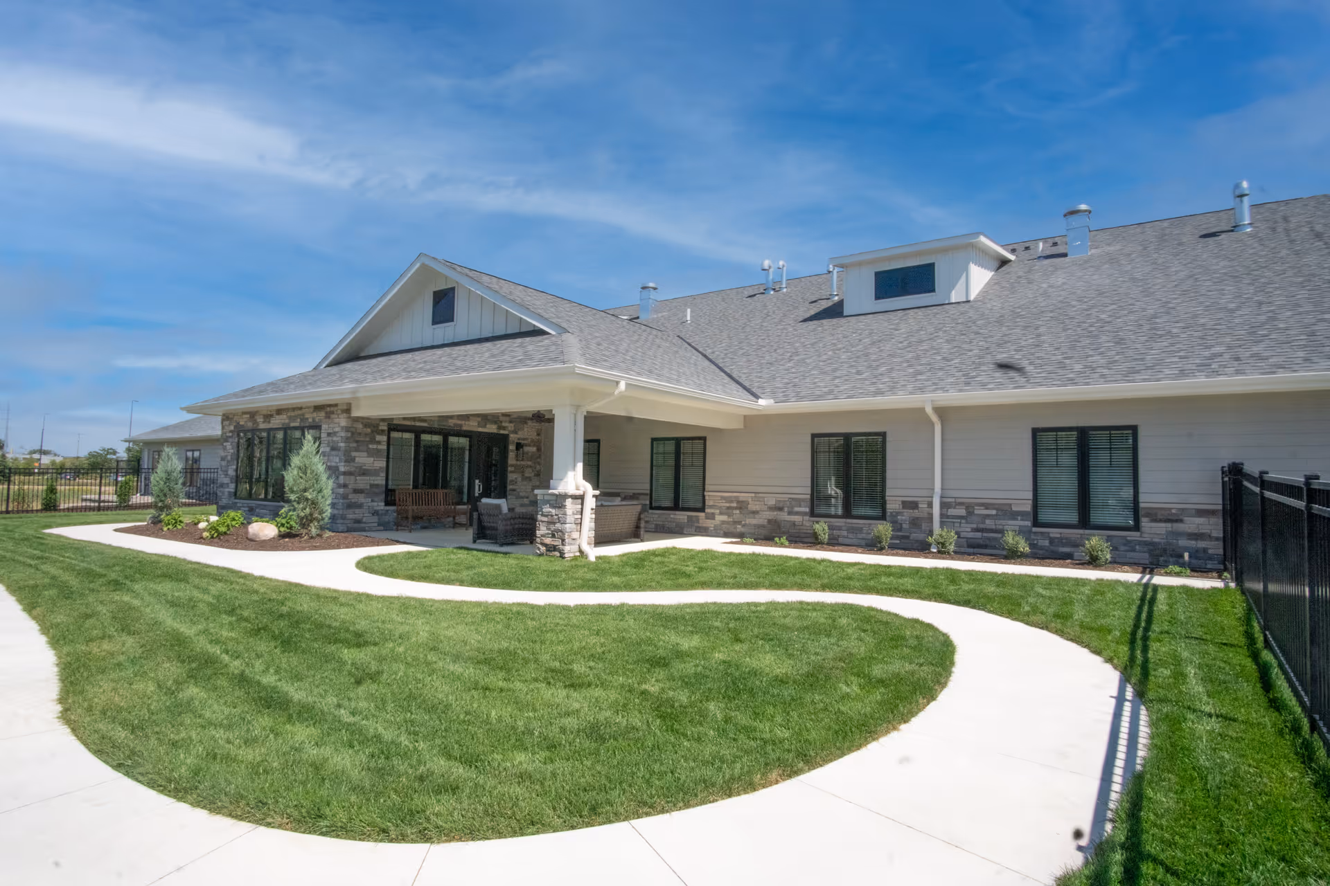 Modern senior living facility exterior with curved path, benches, and manicured lawn.