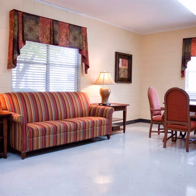 A cozy sitting area with a striped sofa in shades of red, orange, and beige, a wooden side table with a decorative lamp, two red upholstered chairs around a wooden table, and windows with patterned valances letting in natural light.