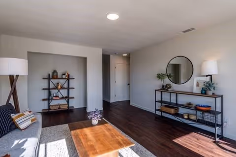 A bright living room with wooden flooring, a gray sofa with cushions, a wooden coffee table, a tall floor lamp, a black metal shelf with decorative items, and a round mirror on the wall above a console table with plants and decor.