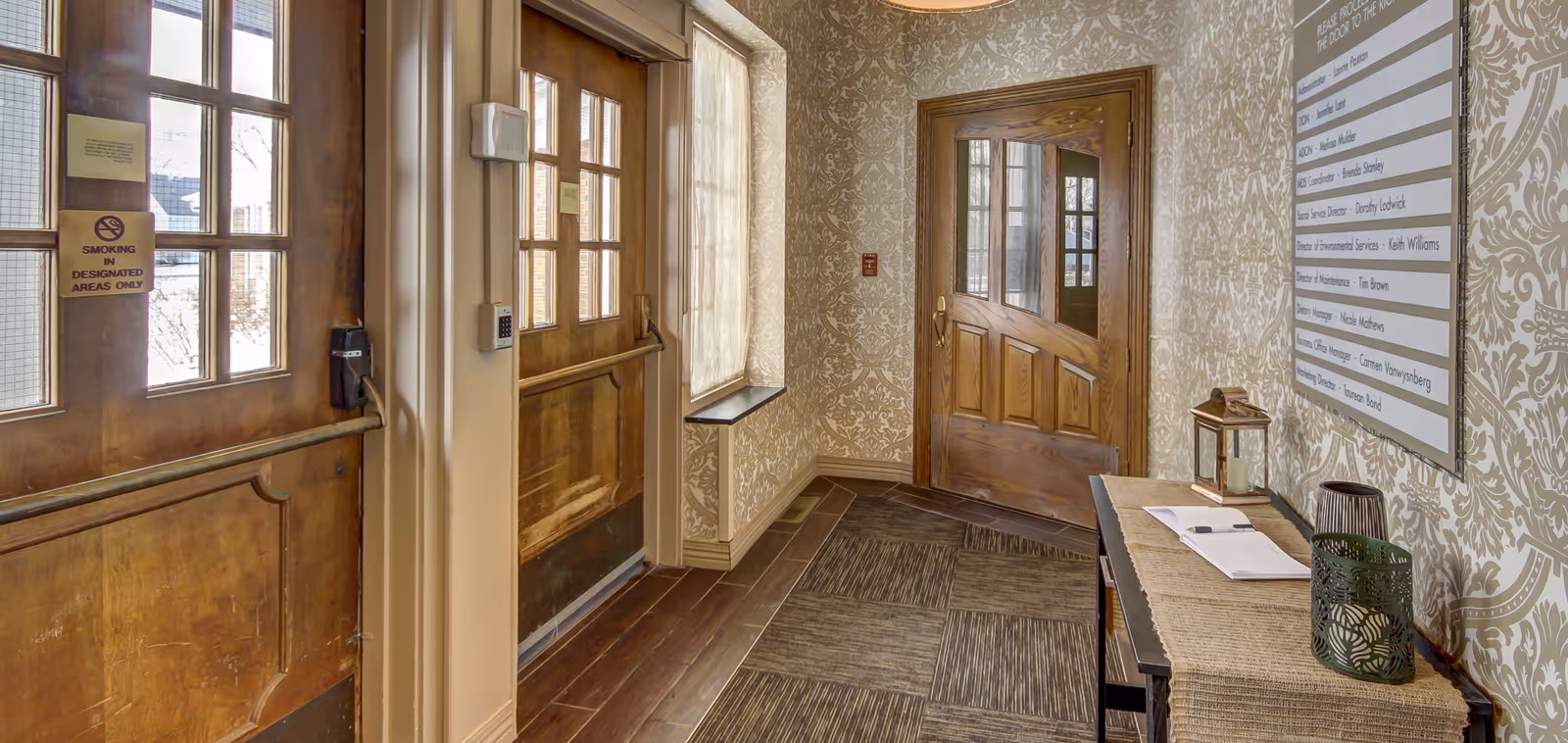 Interior hallway with wooden doors and patterned wallpaper. A small table with decorative items and an open book is placed against the wall. A sign with names and titles is mounted on the wall above the table. The floor has a combination of wood and carpet tiles.