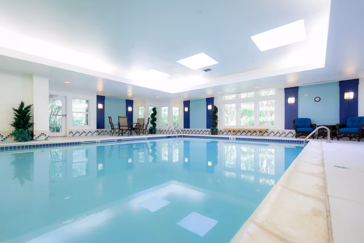 Indoor swimming pool with clear blue water, surrounded by white and blue tiled walls. There are several windows letting in natural light, potted plants, and seating areas with chairs and tables around the pool. The ceiling has recessed lighting and skylights.