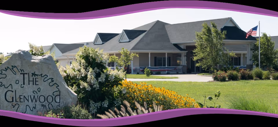Exterior view of The Glenwood Assisted Living of Effingham building with a large rock sign in the foreground surrounded by flowers and greenery, an American flag on a flagpole, and a well-maintained lawn.