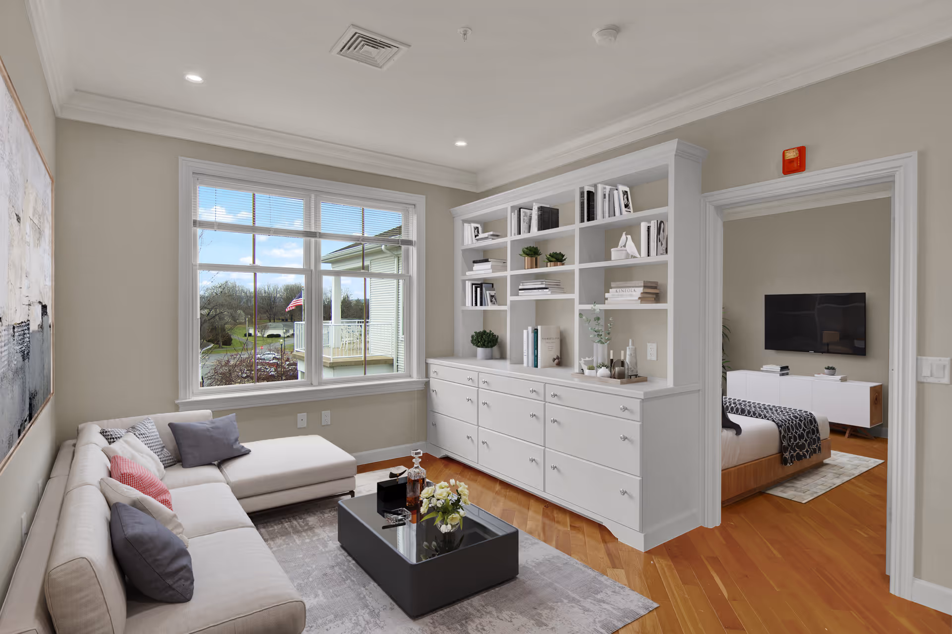 A bright living room with a large window showing an outdoor view with an American flag. The room features a beige sectional sofa with multiple pillows, a black coffee table with decorative items, and a built-in white shelving unit filled with books and plants. Through an open doorway, a bedroom with a bed, a patterned throw, and a wall-mounted TV is visible.
