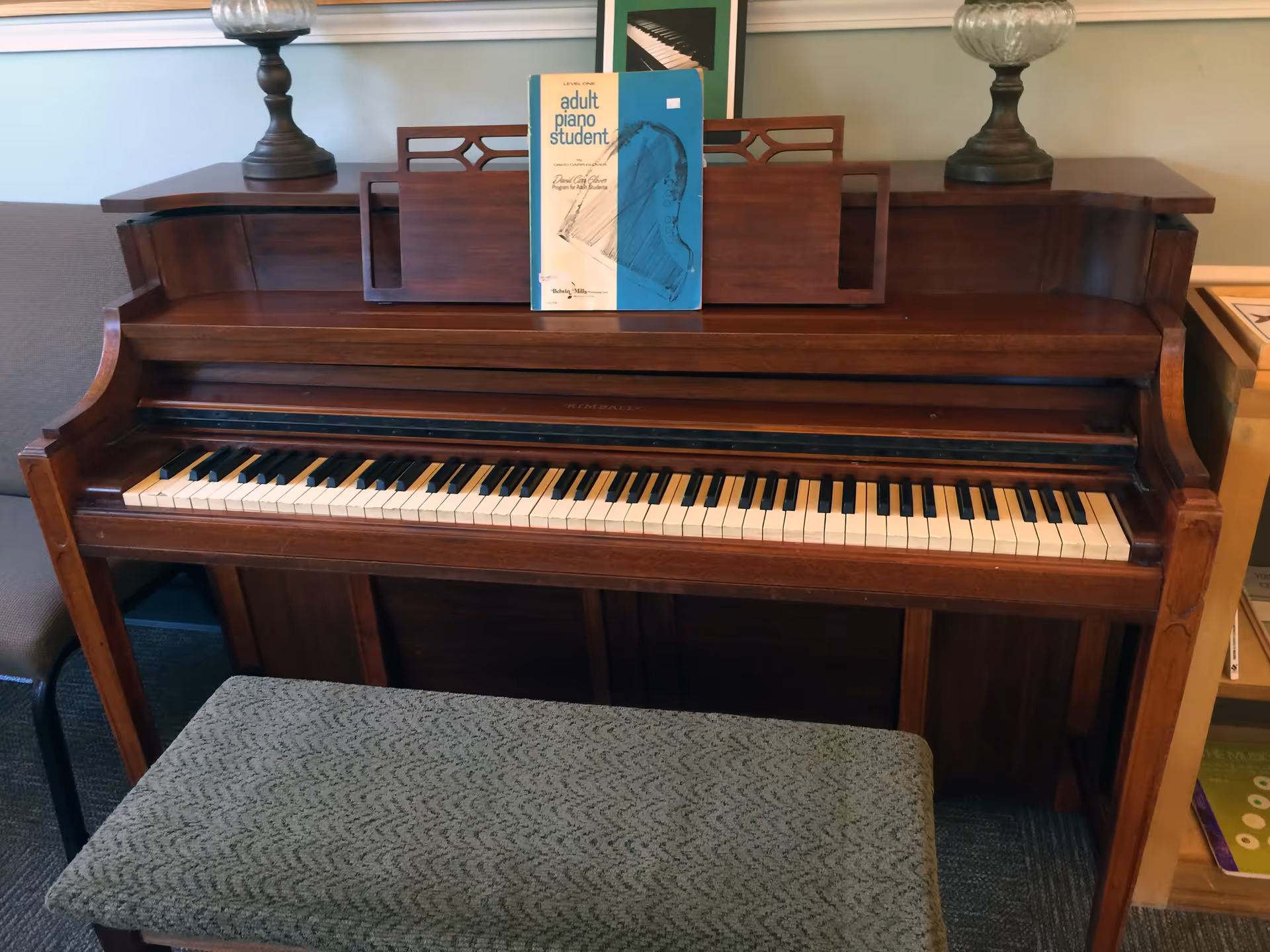 A wooden upright piano with a music book on the stand and a padded bench in front inside a common room.