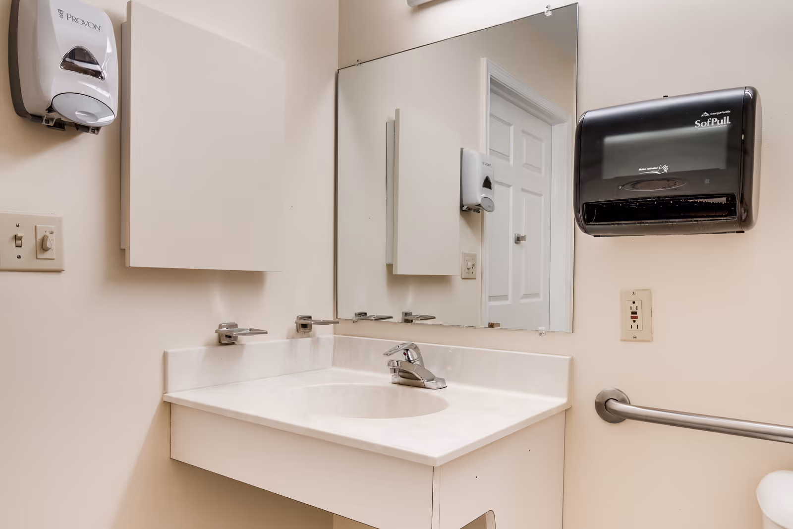 A clean bathroom sink area with a white countertop and a single faucet. Above the sink is a large rectangular mirror reflecting a closed white door. On the left wall, there is a white soap dispenser, and on the right wall, there is a black paper towel dispenser and a metal grab bar. The walls are painted a light beige color.