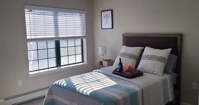 Sunlit bedroom with a neatly made bed, striped bedding, a nightstand with a lamp and tray, and a window with blinds.