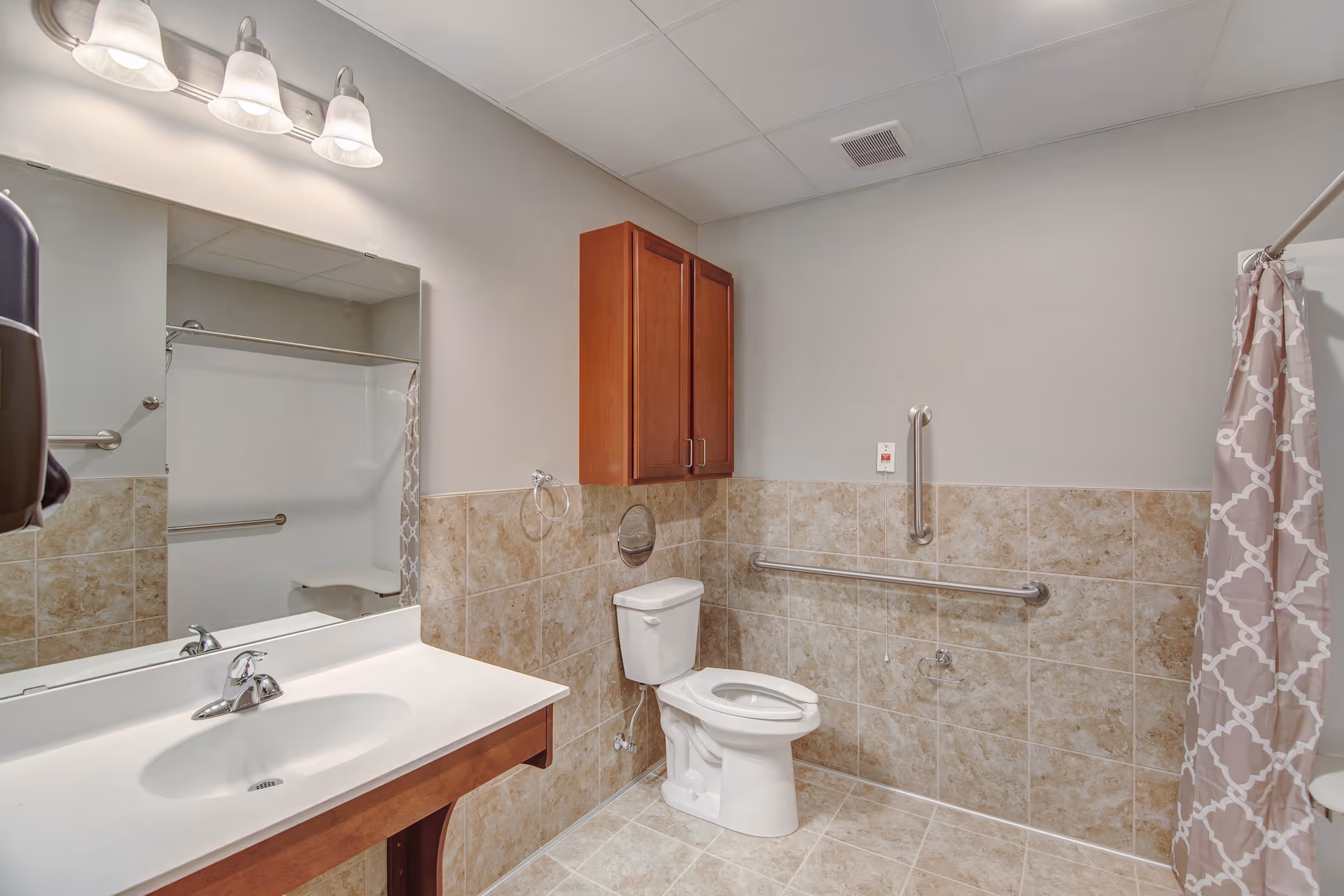 A clean and accessible bathroom with beige tiled walls and floor. The bathroom features a white sink with a chrome faucet, a large mirror above the sink, a wooden cabinet mounted on the wall, a white toilet with grab bars on the wall for support, and a shower area with a patterned shower curtain.
