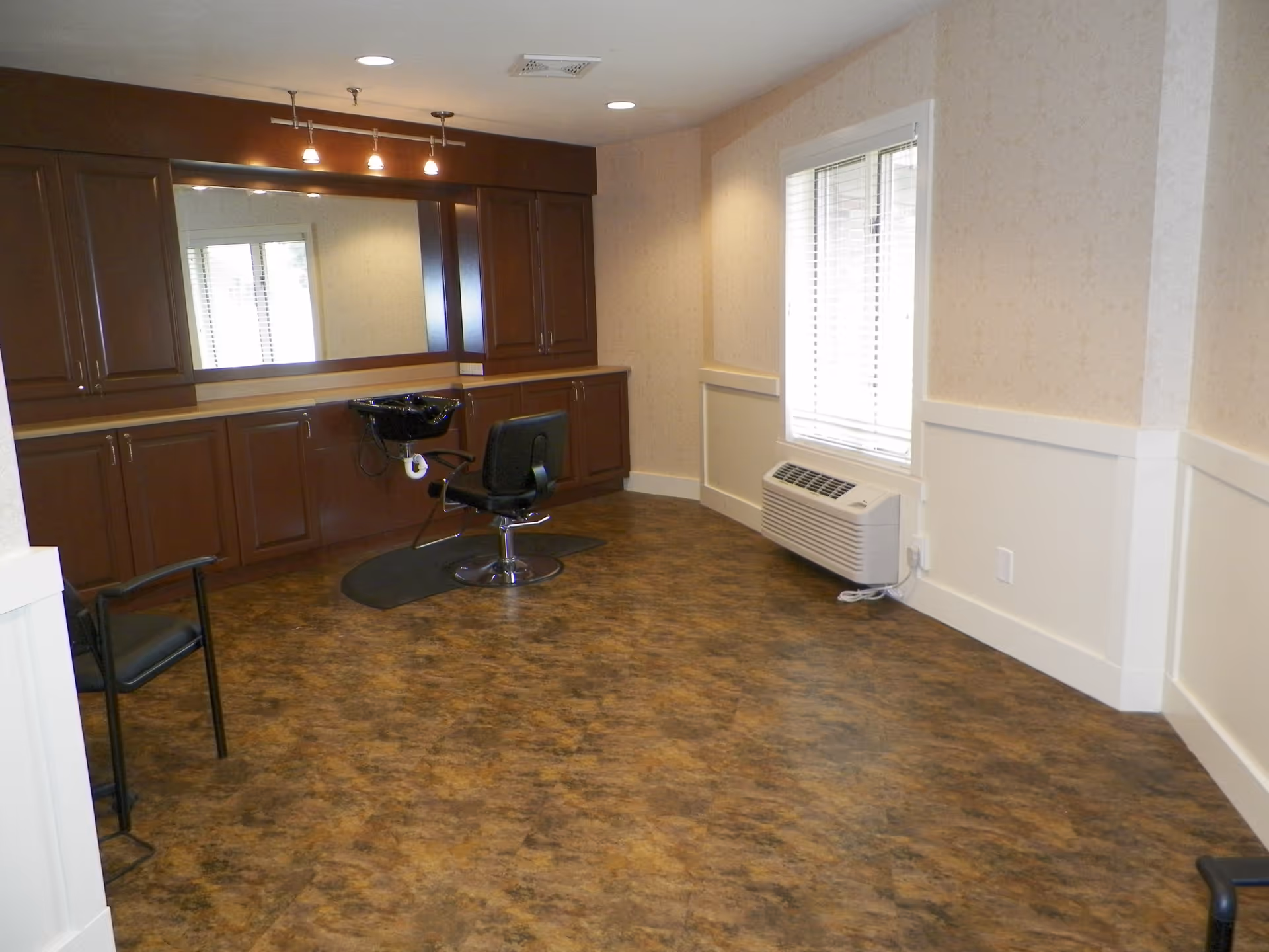 Interior room with a salon chair and a black hair washing sink in front of a large mirror mounted on a wall with wooden cabinets. The room has a window with blinds and a wall-mounted air conditioning unit below it. The floor has a brown patterned surface and the walls are light-colored with white trim.