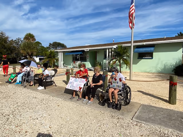 Residents seated outside in front of a single-story light-green assisted living building holding handmade signs beneath an American flag.