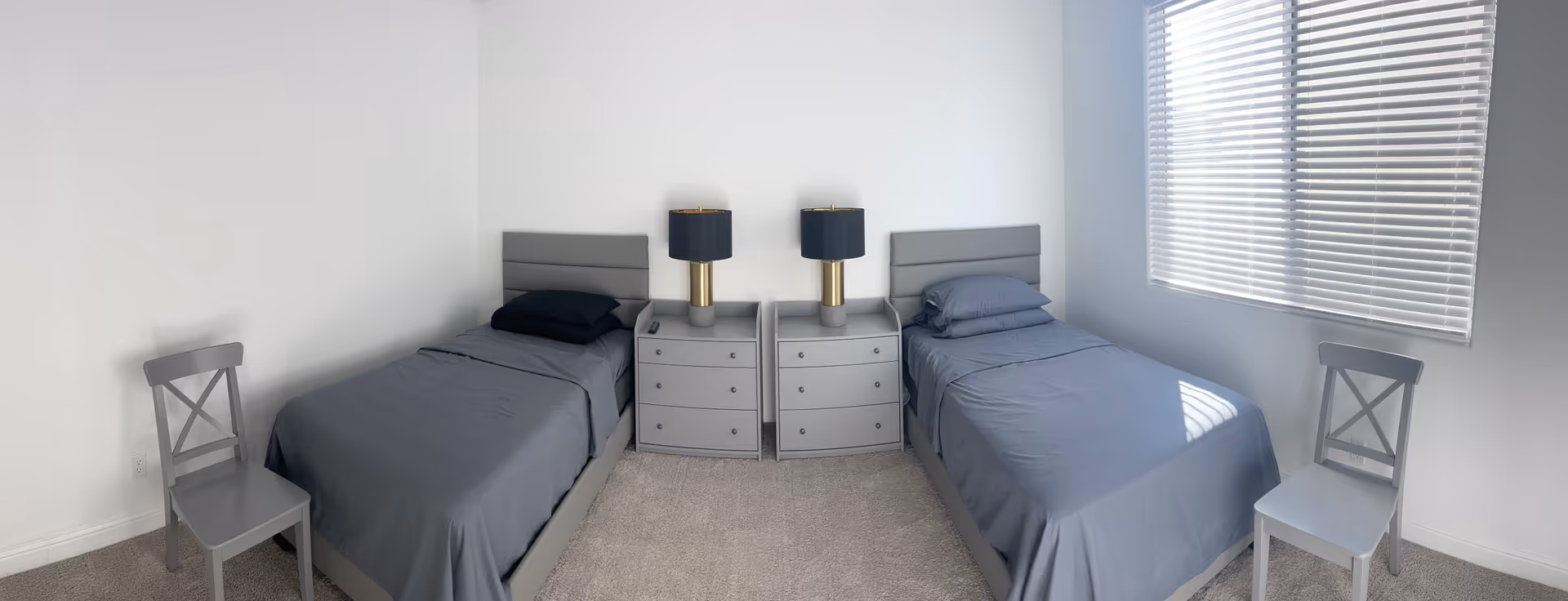 A simple, clean bedroom with two single beds separated by two gray nightstands, each with a black and gold lamp. Each bed has gray bedding and a pillow. There are two gray chairs, one placed near each bed. A window with closed blinds is on the right wall, allowing some natural light into the room. The walls are white and the floor is carpeted in a neutral color.