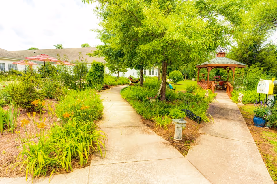 A bright outdoor garden area with a concrete pathway that splits into two directions. The garden is lush with green plants, flowers, and trees. On the right side, there is a wooden gazebo with seating inside. In the background, a building with a sloped roof and windows is visible. There are also red umbrellas over tables near the building.