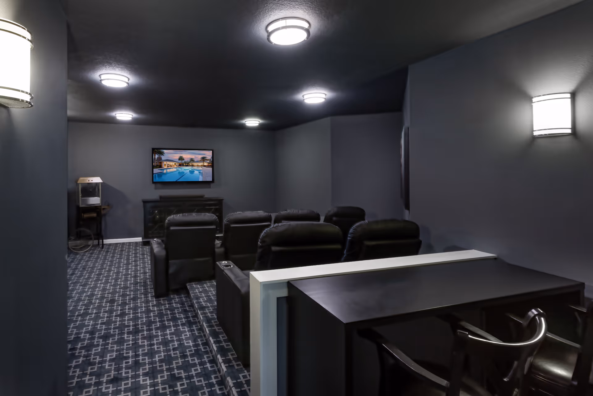 A small home theater room with dark walls and ceiling, featuring six black leather recliner chairs arranged in two rows facing a wall-mounted flat-screen TV displaying a pool scene. The room has patterned carpet flooring, wall-mounted lights, and a popcorn machine in the corner.