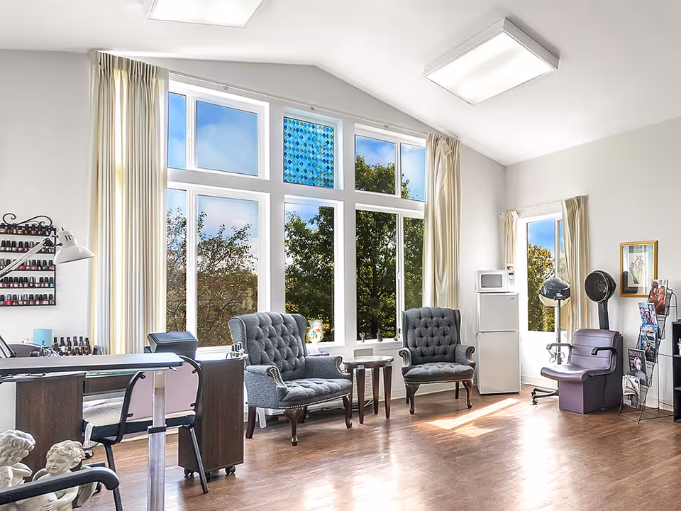 Bright room with large windows showing trees outside, featuring two gray tufted armchairs with a small table between them, a nail polish rack on the wall, a manicure table with chairs, a white refrigerator and microwave, a purple salon chair with a hair dryer, and a magazine rack.