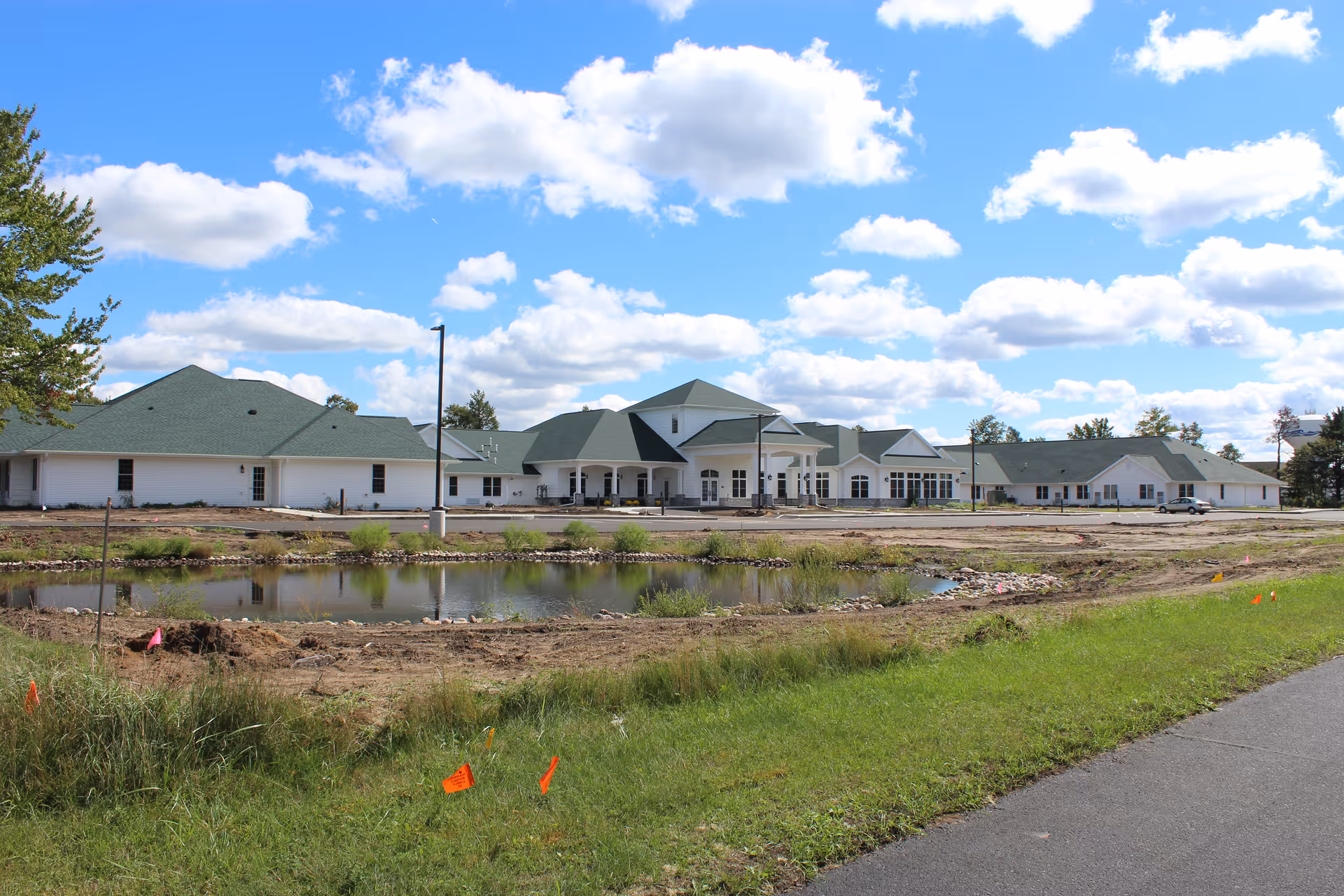Exterior view of a large single-story building with green roofs and white walls under a blue sky with scattered clouds. In front of the building is a small pond surrounded by grass and some construction markers, with a paved road in the foreground.