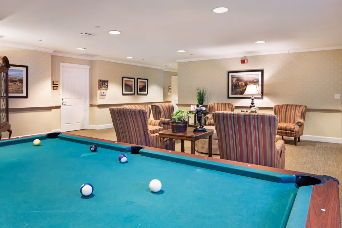 A cozy indoor common area featuring a pool table in the foreground with several billiard balls on it. In the background, there are four striped armchairs arranged around a wooden coffee table with plants and decorative items. The walls are adorned with framed landscape paintings, and the room is softly lit with recessed ceiling lights and a table lamp.