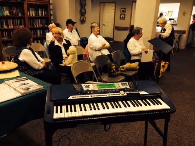 A group of elderly people seated on folding chairs in a room, some holding stuffed animals. In the foreground, there is an electronic keyboard on a stand. The room has bookshelves, framed pictures on the walls, and a carpeted floor.