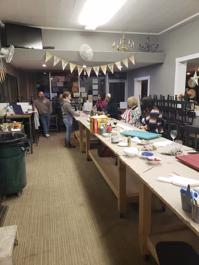 A group of people gathered around a long table in a room with art supplies and paint materials. The room has a banner hanging from the ceiling and several chairs stacked against the wall. The setting appears to be a communal or activity room with a casual atmosphere.