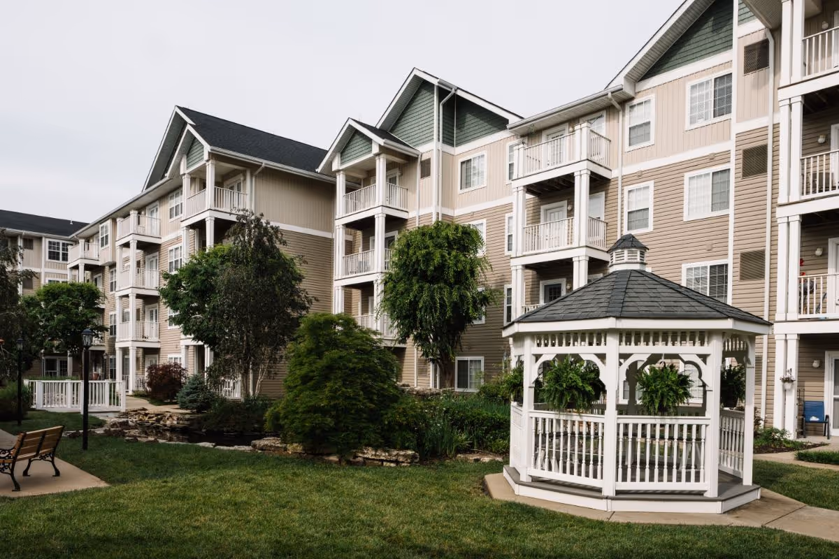 Exterior courtyard of a multi-story senior living building with a white gazebo, landscaped greenery, and balconies.
