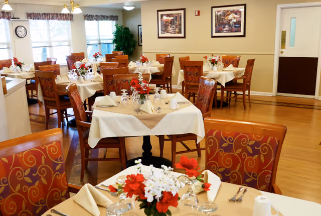 A bright dining room with multiple tables set with white tablecloths, floral centerpieces, and patterned wooden chairs.