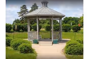 Wooden gazebo with a shingled roof in a landscaped garden with a paved path, benches, shrubs, and trees.