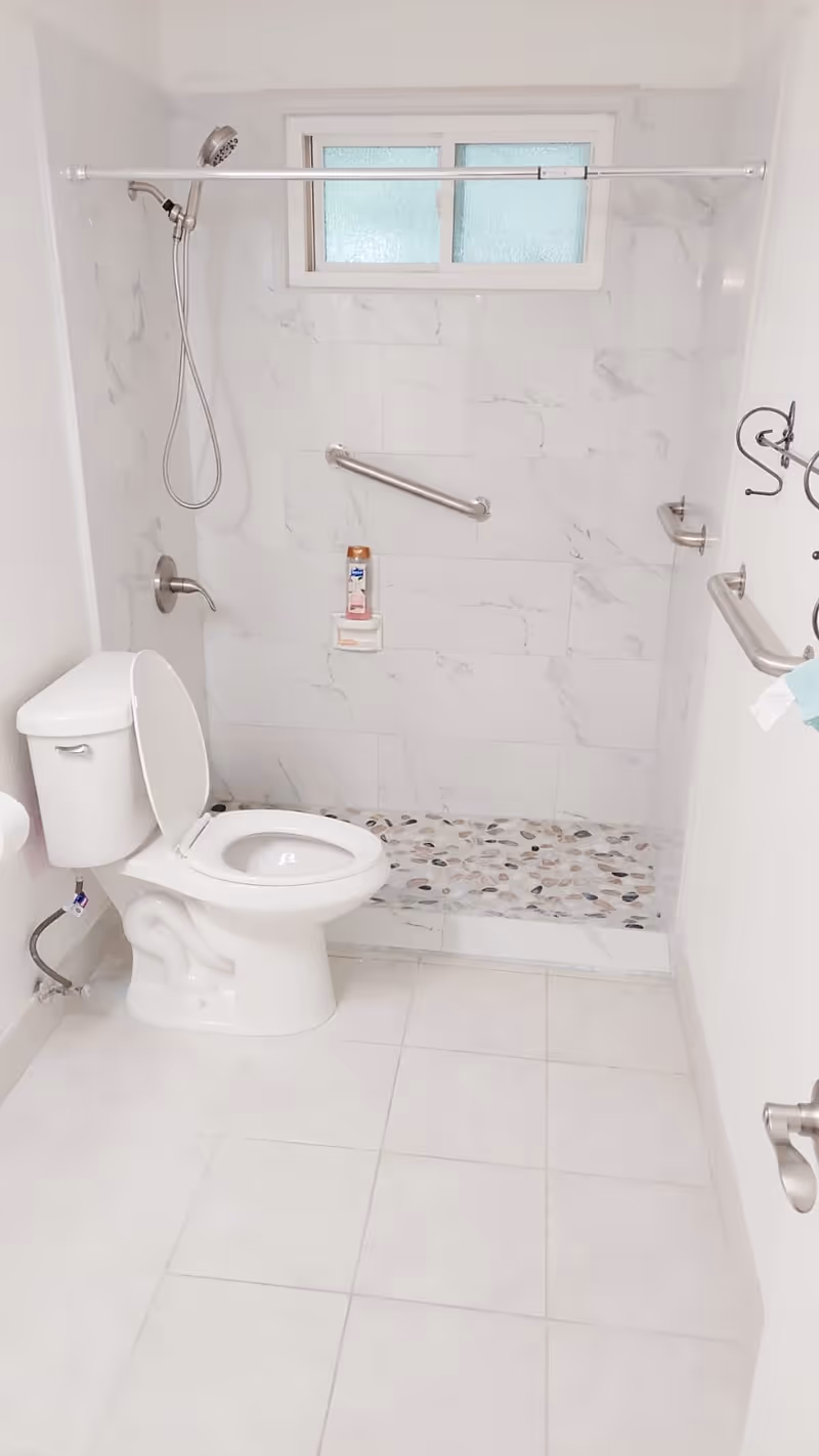 A clean, white bathroom with a toilet and an open shower area. The shower has a handheld showerhead, a grab bar, and a small window above. The shower floor is tiled with pebble-like stones, and the walls have white marble-patterned tiles. There are also towel hooks and a grab bar on the right wall.