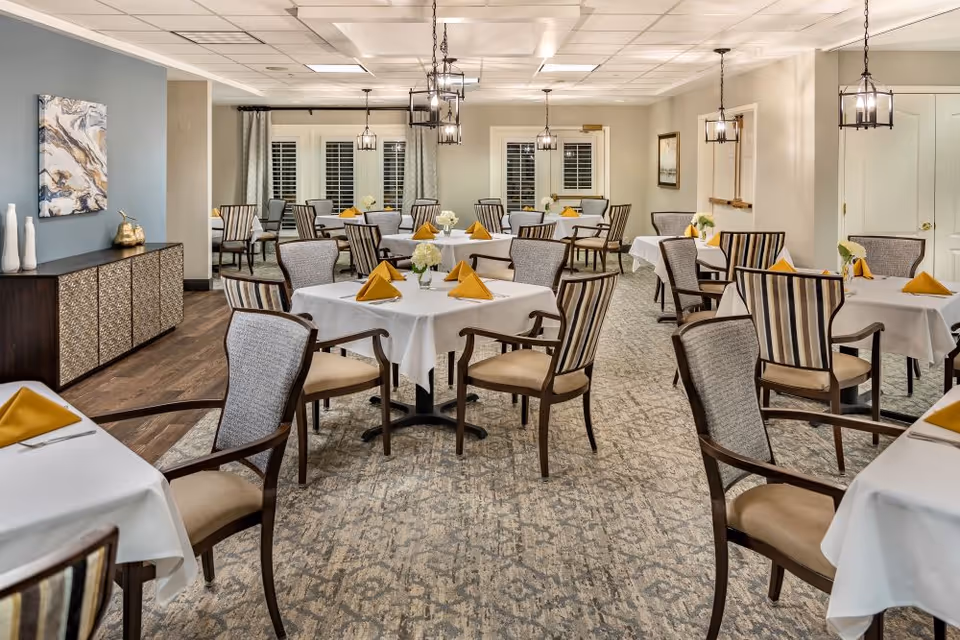A well-lit dining room with multiple tables covered in white tablecloths, each set with yellow folded napkins and small floral centerpieces. The room features wooden chairs with cushioned seats and backs, a patterned carpet, and modern hanging light fixtures. There is a sideboard against one wall with decorative items and a piece of abstract art above it.
