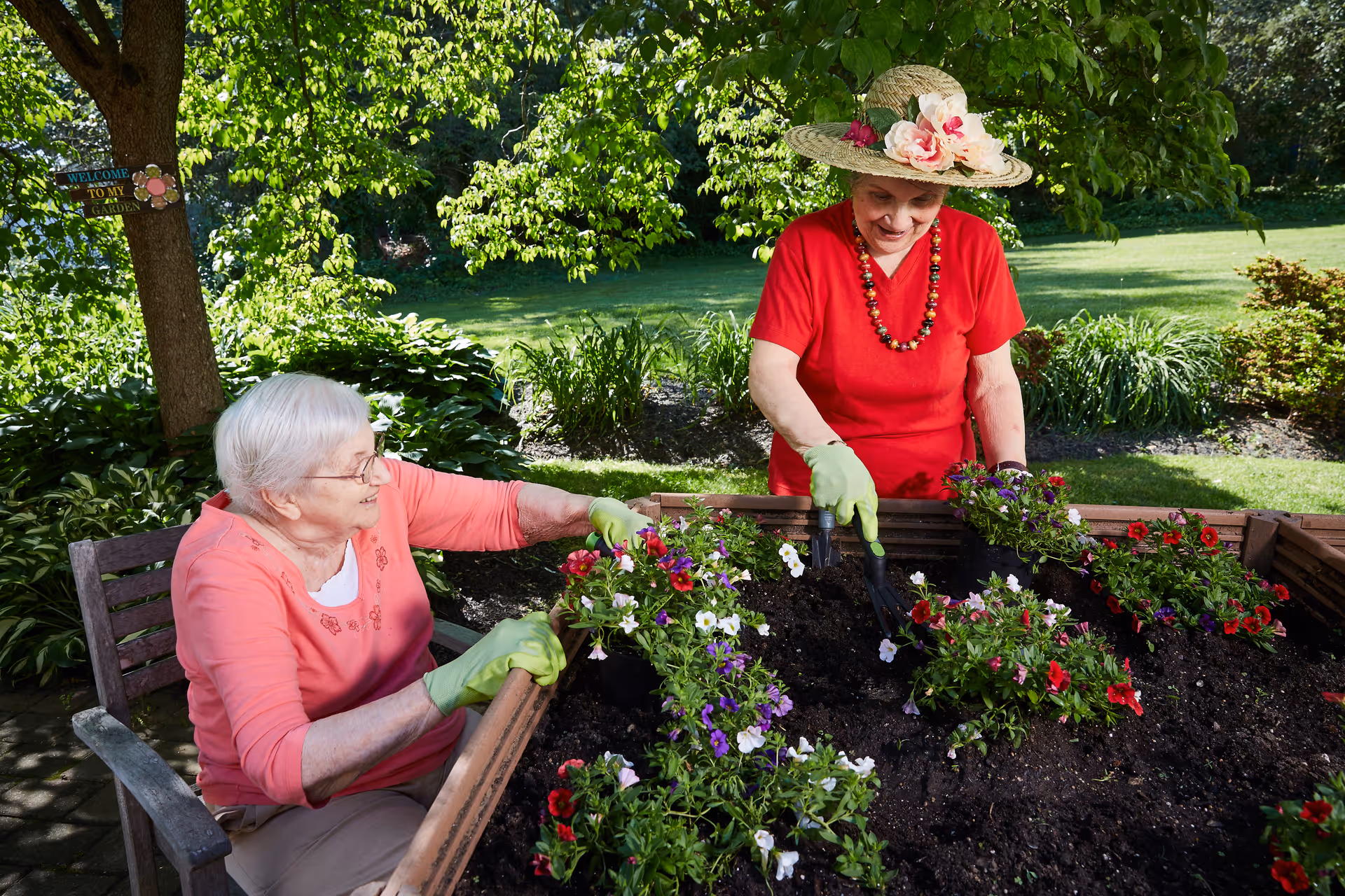 Two elderly women gardening outdoors in a raised flower bed. One woman is seated wearing a pink shirt and green gloves, while the other is standing wearing a red shirt, green gloves, and a wide-brimmed hat decorated with flowers. They are planting colorful flowers in a sunny garden with green trees and shrubs in the background.