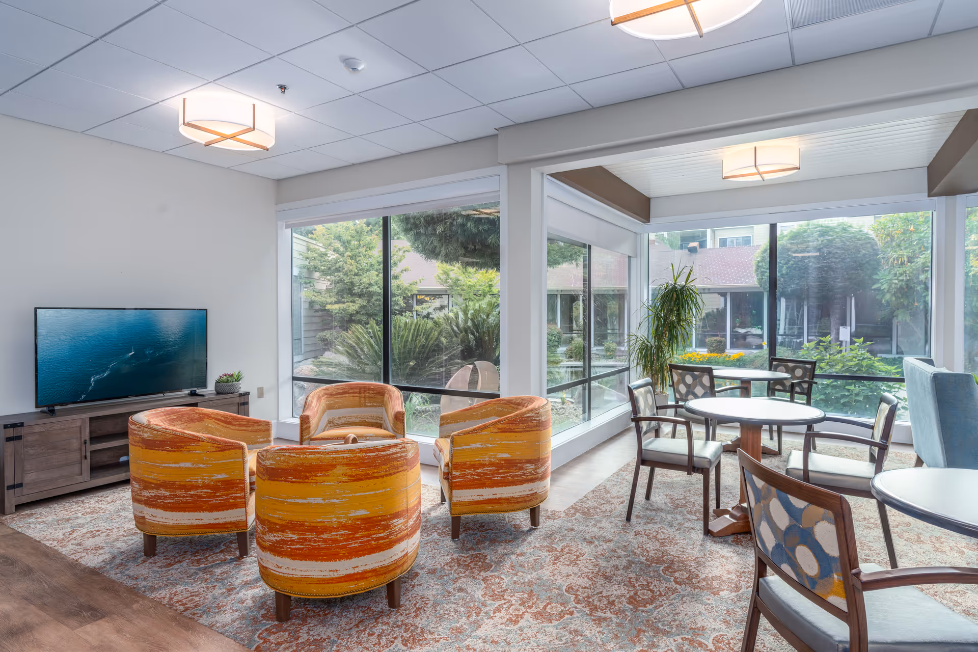 A bright and cozy common area with large windows overlooking a garden. The room features four orange and yellow patterned armchairs arranged around a small table on a decorative rug. There is a flat-screen TV on a wooden stand to the left and several round tables with chairs near the windows. The ceiling has modern light fixtures.