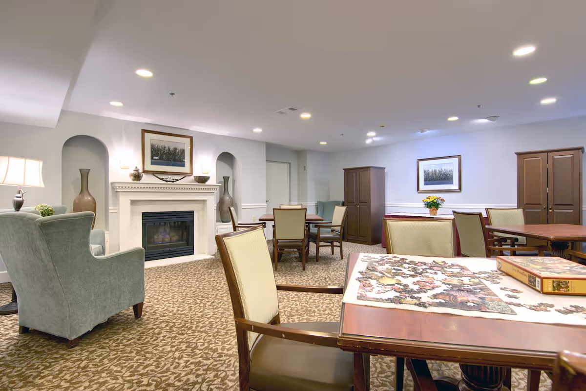 A cozy senior living community common room with a fireplace, armchairs, tables, and chairs. One table has a partially completed jigsaw puzzle and a puzzle box on it. The room is carpeted with a patterned design, and the walls are decorated with framed artwork and large vases. There are cabinets and a lamp providing soft lighting.