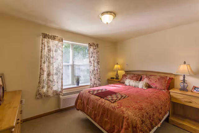 A cozy bedroom with a large bed covered in a red and gold patterned comforter and pillows. There are two wooden nightstands on either side of the bed, each with a lamp. A window with floral curtains lets in natural light, and a wooden dresser is visible on the left side of the room.