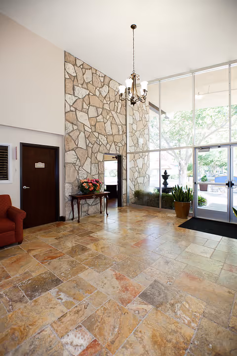 Sunlit building lobby with a stone accent wall, tiled floor, chandelier, glass entrance doors, and a small table with flowers.