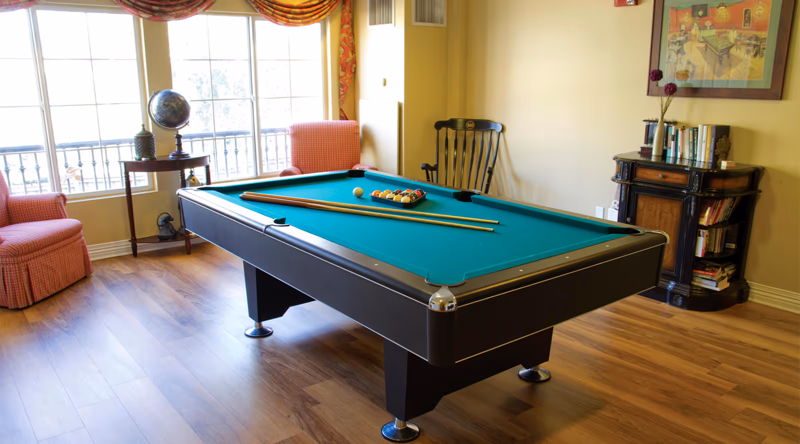 A billiards table in a sunny common room with chairs, a small table holding a globe by the window, and a bookshelf against the wall.
