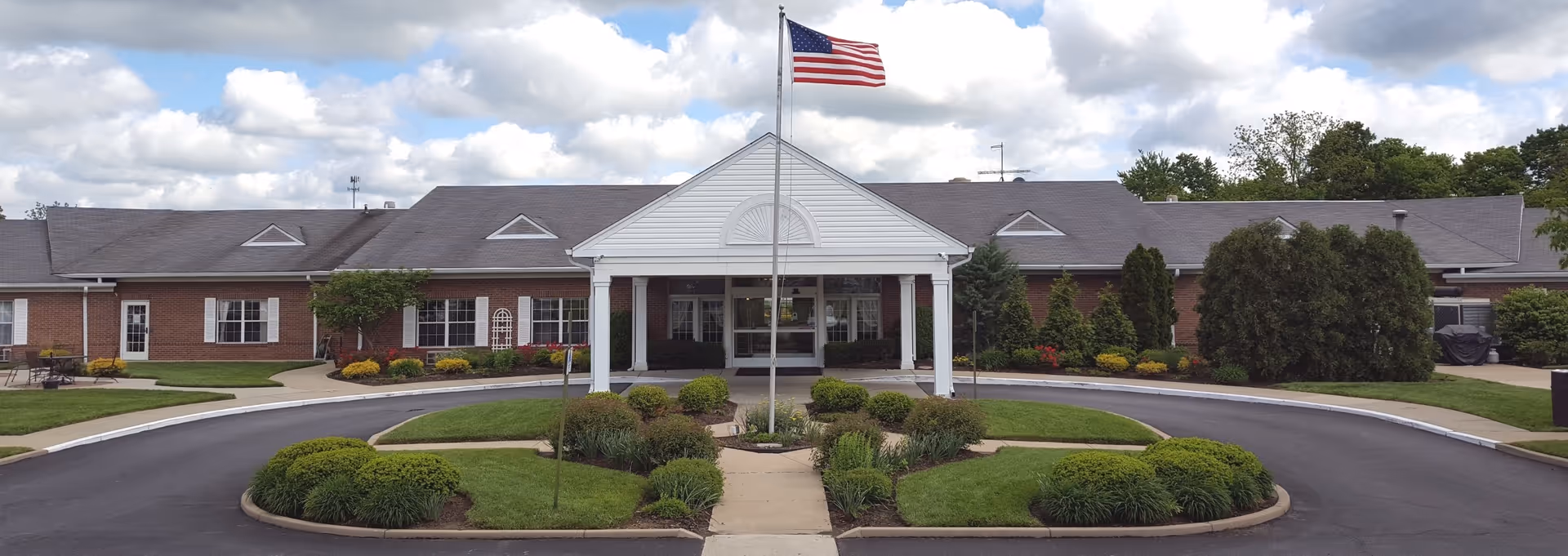 Front exterior view of The Anderson Nursing and Rehabilitation Center, a single-story brick building with a covered entrance supported by white columns, an American flag flying on a flagpole in front, and landscaped greenery including bushes and grass around a circular driveway.