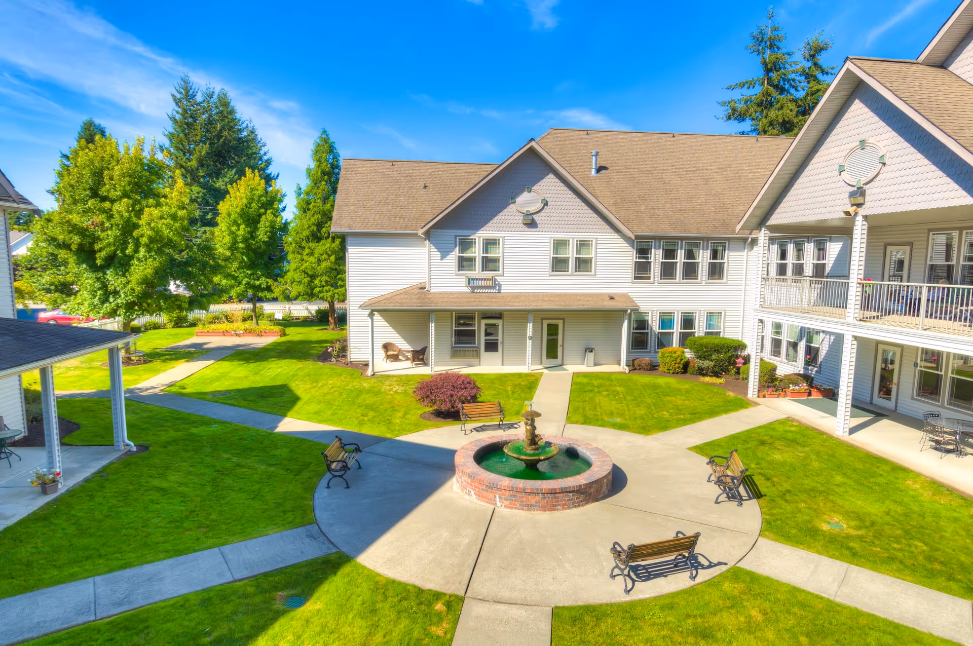 A bright outdoor courtyard area of a senior living facility with a circular concrete pathway surrounding a brick fountain in the center. Several benches are placed around the fountain. The courtyard is surrounded by well-maintained green lawns, trees, and two-story buildings with white siding and multiple windows under a clear blue sky.