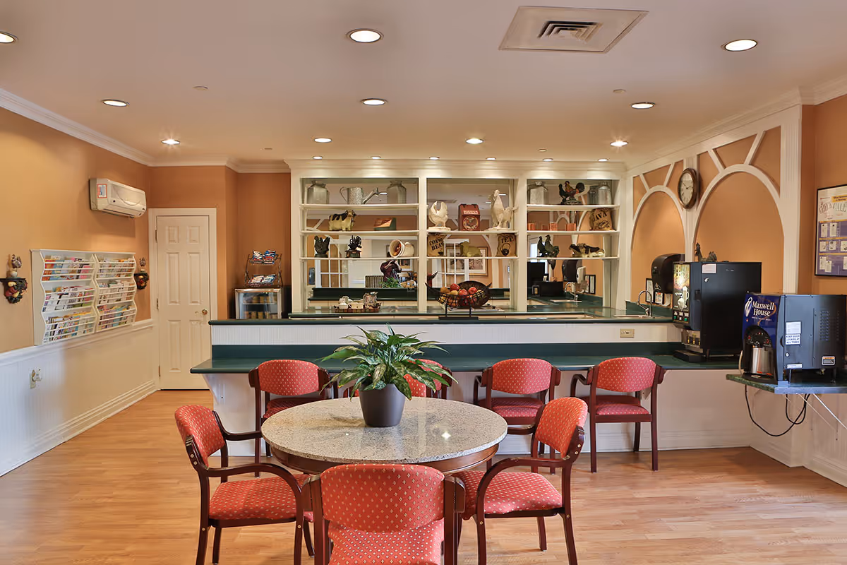 A cozy common area with a round table surrounded by six red cushioned chairs. A potted plant is centered on the table. Behind the table is a counter with green countertops and decorative shelves displaying various ornaments. To the right, there are coffee machines and a clock on the wall. The room has warm beige walls, wooden flooring, and recessed ceiling lights.