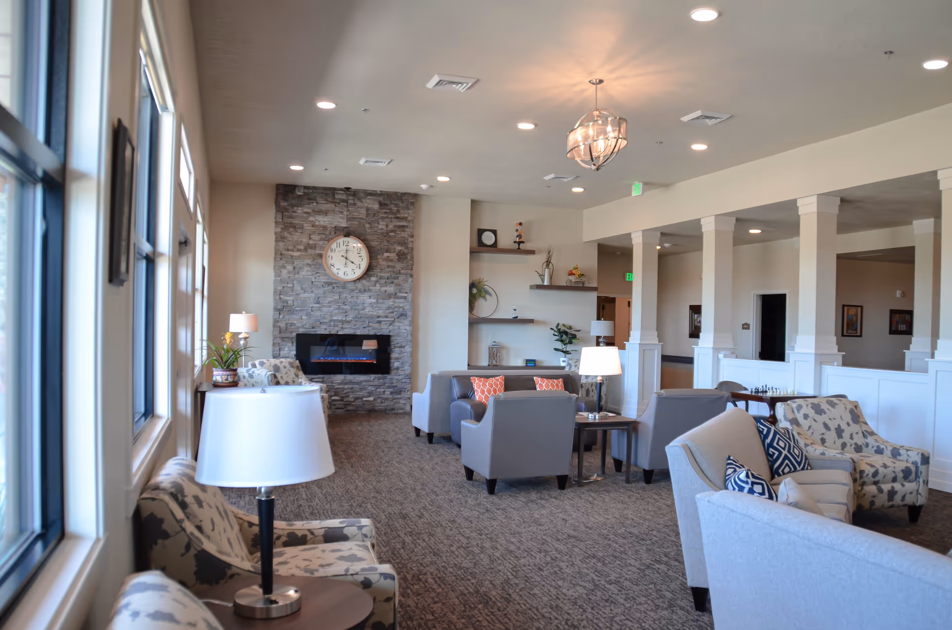 A spacious senior living facility common area with multiple seating arrangements including armchairs and sofas. The room features a stone fireplace with a clock above it, large windows on the left side letting in natural light, decorative shelves with plants and ornaments, and ceiling lights including a chandelier. The carpeted floor and neutral wall colors create a warm and inviting atmosphere.